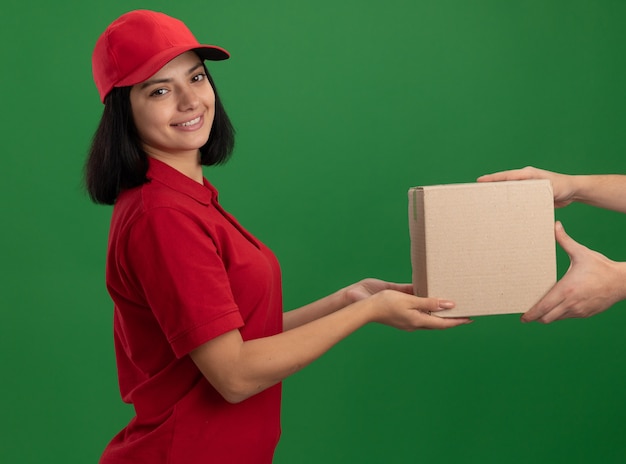 Jong leveringsmeisje in rood uniform en glb die kartondoos geven aan een klant vriendelijk glimlachend status over groene muur