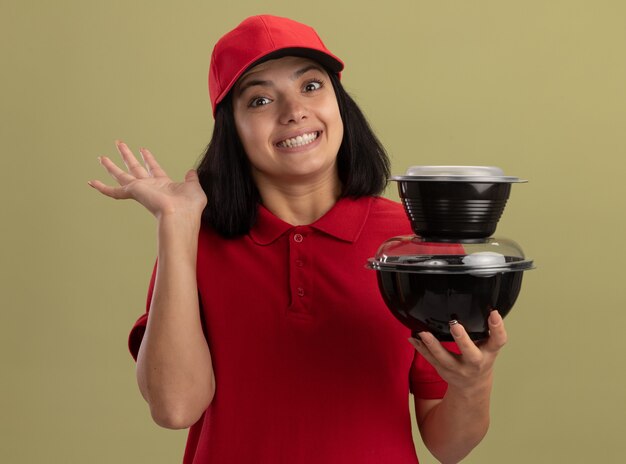 Jong leveringsmeisje in rood uniform en GLB die voedselpakketten glimlachen die met hand golven die zich over lichte muur bevinden