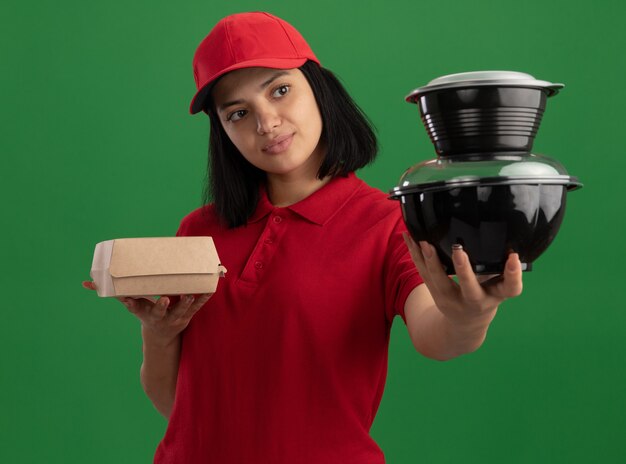 Jong leveringsmeisje in rood uniform en pet met voedselpakketten die zelfverzekerd over groene muur kijken