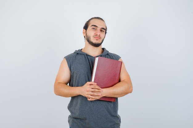 Jong mannelijk boek terwijl poseren in mouwloze hoodie en er zelfverzekerd uitzien. vooraanzicht.