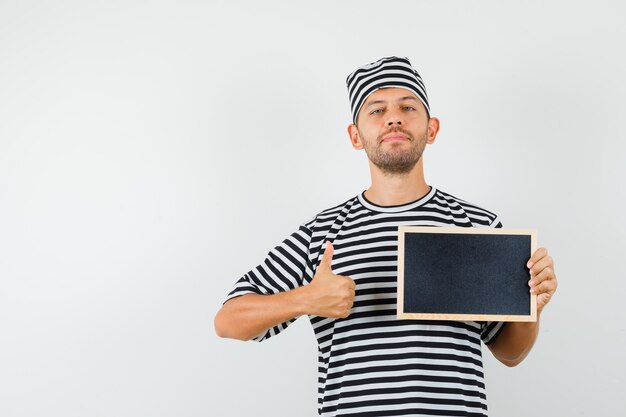 Jong mannelijk holdingsbord dat duim in gestreepte t-shirthoed toont en zelfverzekerd kijkt