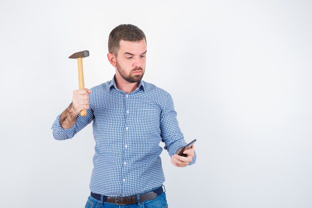 Jong mannetje dat beweert mobiele telefoon te slaan met een hamer in overhemd, spijkerbroek en ernstig, vooraanzicht kijkt.