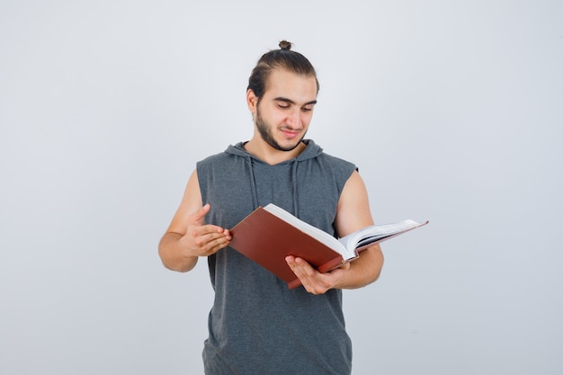 Jong mannetje dat boek in mouwloze hoodie bekijkt en gericht kijkt. vooraanzicht.