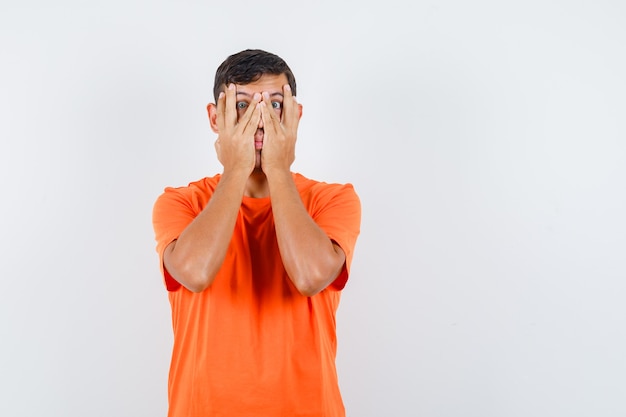 Jong mannetje dat door vingers in oranje t-shirt kijkt en bang kijkt