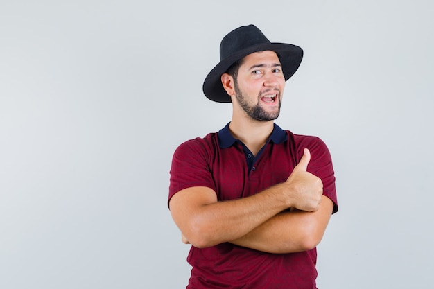 Jong mannetje dat duim in t-shirt, hoed toont en positief kijkt. vooraanzicht.