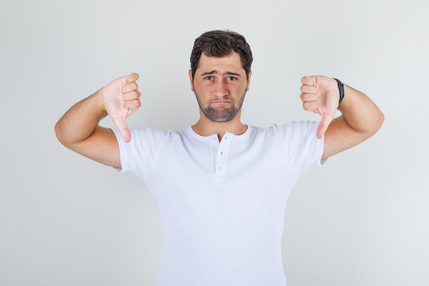 Jong mannetje dat duimen neer in wit t-shirt toont en verdrietig kijkt