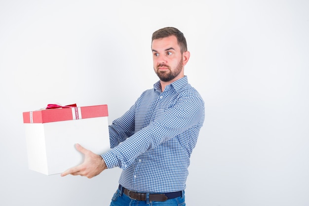 Jong mannetje dat het geven van gebaar toont terwijl giftdoos in overhemd, spijkerbroek en op zoek zelfverzekerd, vooraanzicht.