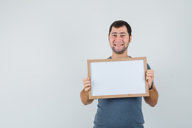 Jong mannetje dat in grijs t-shirt leeg frame houdt en vrolijk kijkt