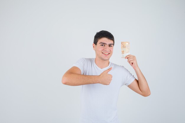 Jong mannetje dat in t-shirt eurobanknote houdt, duim toont en tevreden, vooraanzicht kijkt.