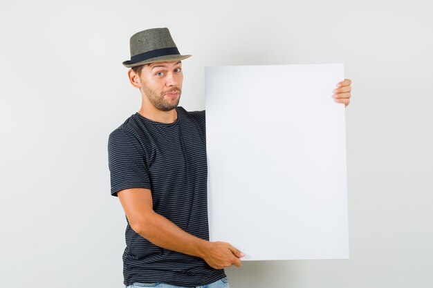 Gratis foto jong mannetje dat leeg canvas in de hoed van t-shirtjeans houdt en zelfverzekerd kijkt