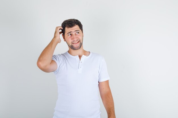 Jong mannetje dat met hand op hoofd in wit t-shirt omhoog kijkt en verlegen kijkt