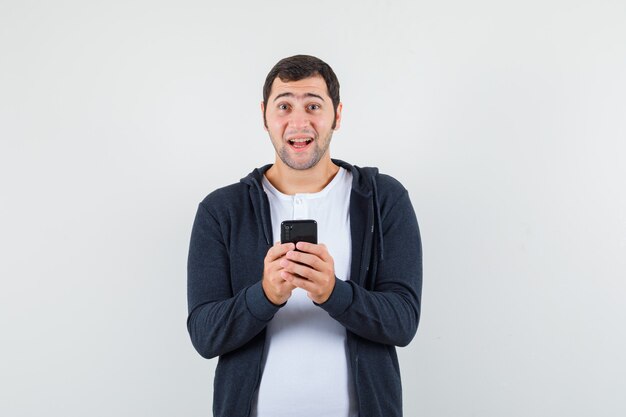 Jong mannetje dat mobiele telefoon in t-shirt, jasje houdt en blij kijkt. vooraanzicht.