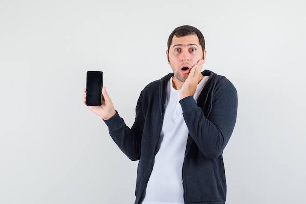 Jong mannetje dat mobiele telefoon in t-shirt, jasje houdt en verbaasd kijkt. vooraanzicht.