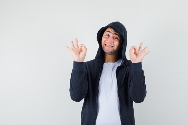 Jong mannetje dat ok gebaar in t-shirt, jasje toont en vrolijk, vooraanzicht kijkt.