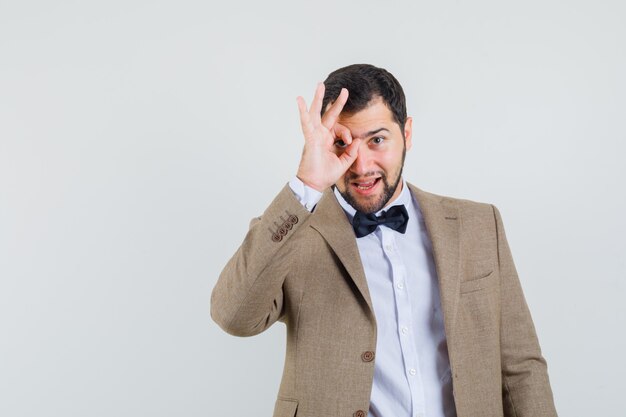 Jong mannetje dat ok teken op oog in kostuum toont en zelfverzekerd kijkt. vooraanzicht.