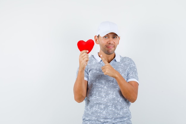 Gratis foto jong mannetje dat op rood hart in t-shirt en pet richt en vrolijk kijkt