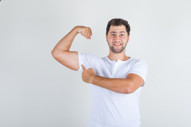 Jong mannetje dat spier met vinger in wit t-shirt toont en sterk kijkt