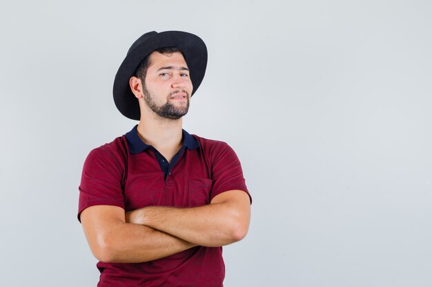 Jong mannetje dat zich met gekruiste wapens in t-shirt, hoed bevindt en zelfvoldaan kijkt. vooraanzicht.