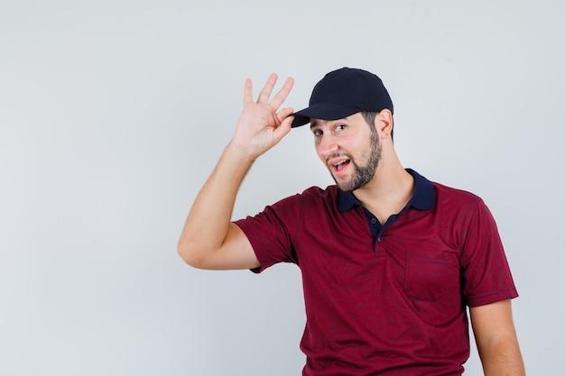 Jong mannetje dat zijn pet vasthoudt terwijl hij naar de camera in rood t-shirt, zwarte pet kijkt en vrolijk kijkt. vooraanzicht.