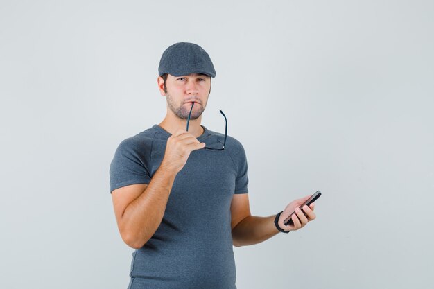 Jong mannetje in grijs t-shirtglb die glazen bijten die mobiele telefoon houden en peinzend kijken