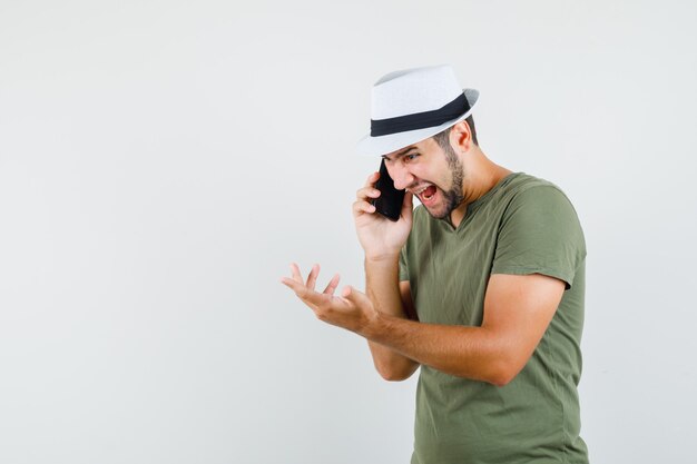 Jong mannetje in groen t-shirt en hoed die op mobiele telefoon spreken en agressief kijken