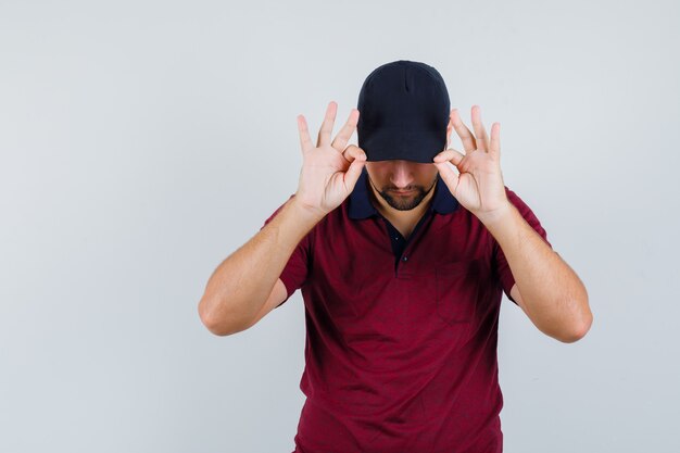 Jong mannetje in rood t-shirt, zwarte pet die pet met vingers aanraakt en er verborgen, vooraanzicht uitziet.