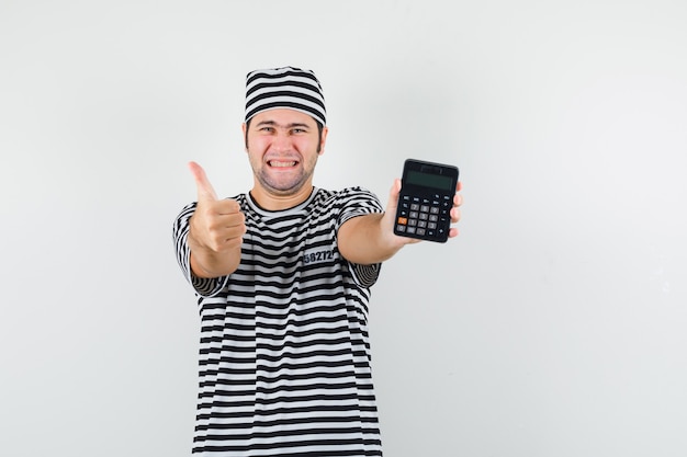 Jong mannetje in t-shirt, hoed die calculator met omhoog duim toont en heel, vooraanzicht kijkt.