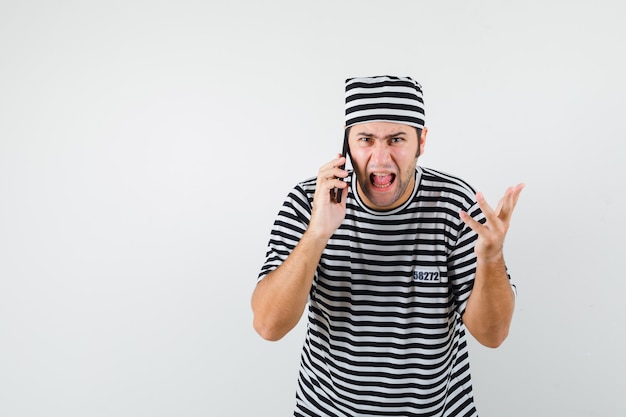 Jong mannetje in t-shirt, hoed die op mobiele telefoon spreekt en zenuwachtig, vooraanzicht kijkt.