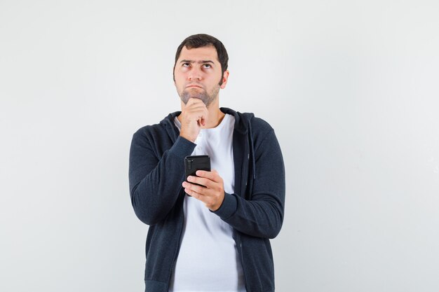 Jong mannetje in t-shirt, jasje die mobiele telefoon houdt en peinzend, vooraanzicht kijkt.