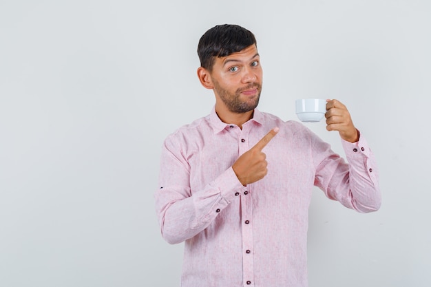 Jong mannetje wijzend op kopje thee en lachend in roze shirt vooraanzicht.