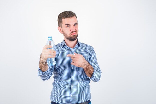 Jong mannetje wijzend op plastic waterfles in shirt, spijkerbroek en kijkt zelfverzekerd, vooraanzicht.