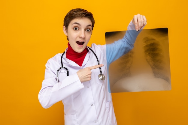 Jong meisje arts in witte jas met stethoscoop met röntgenfoto van longen wijzend met wijsvinger naar voren kijkend blij en verrast over oranje muur