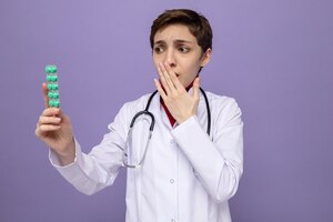 Jong meisje arts in witte jas met stethoscoop om nek met blaar met pillen kijken ernaar bezorgd coning mond met hand staande op paars