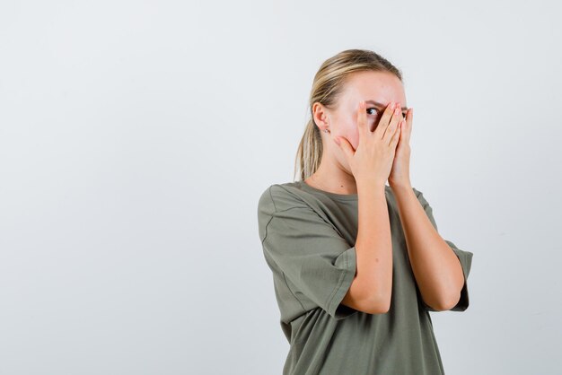 Jong meisje bedekt haar gezicht met haar handen en kijkt bang met haar linkeroog naar de camera op witte achtergrond