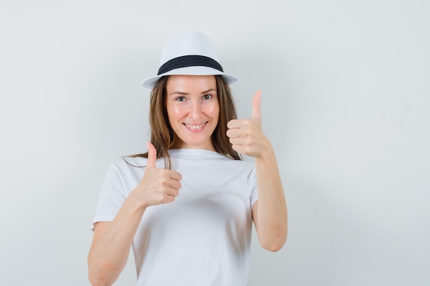 Jong meisje dat dubbele duimen in wit t-shirt, hoed toont en blij, vooraanzicht kijkt.