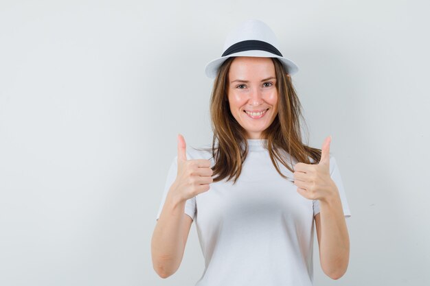 Jong meisje dat dubbele duimen toont in wit t-shirt, hoed en vrolijk, vooraanzicht kijkt.
