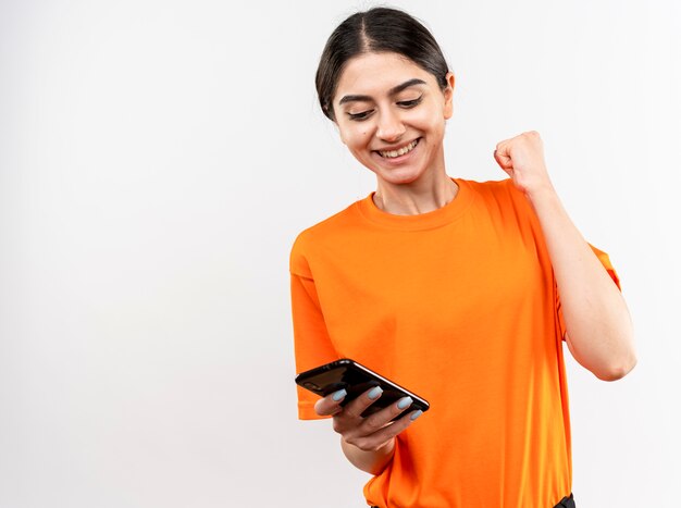 Jong meisje dat een oranje t-shirt draagt dat smartphone met gebalde vuist houdt, blij en opgewonden glimlachend vrolijk verheugd over haar succes staande over witte muur