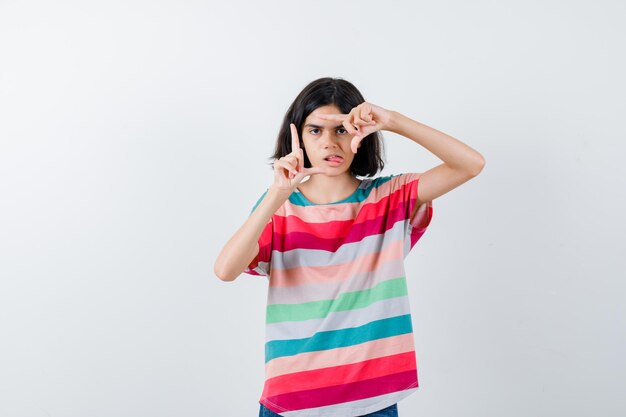 Jong meisje dat framegebaar toont, tong uitsteekt in kleurrijk gestreept t-shirt en er schattig uitziet, vooraanzicht.