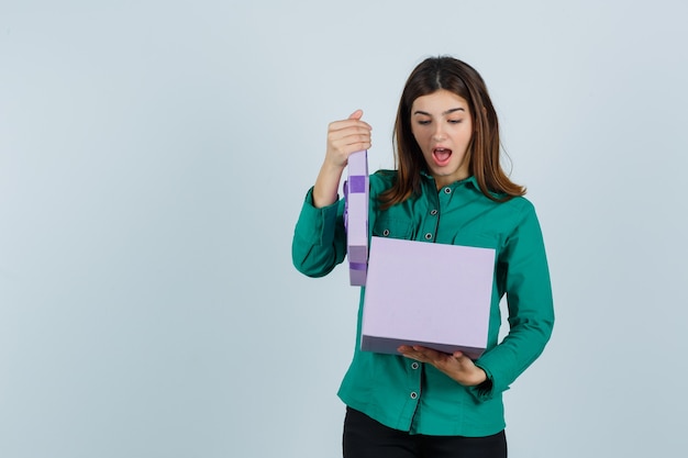 Jong meisje dat giftdoos in groene blouse, zwarte broek probeert te openen en verbaasd kijkt. vooraanzicht.