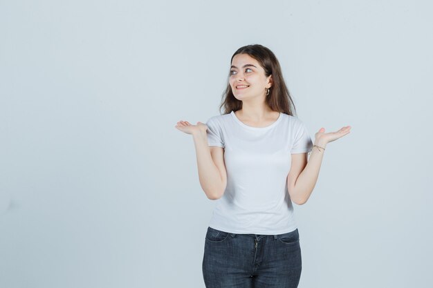 Jong meisje dat handpalmen opzij in t-shirt, jeans uitspreidt en mooi, vooraanzicht kijkt.