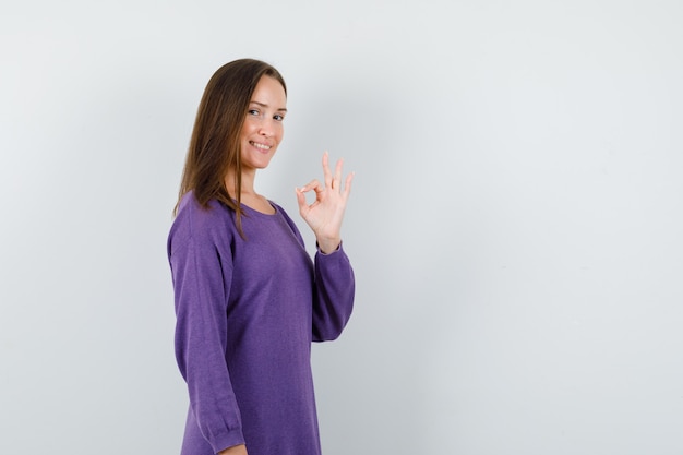 Jong meisje dat ok gebaar in violet overhemd toont en vrolijk, vooraanzicht kijkt.