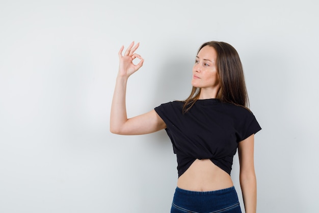 Jong meisje dat ok gebaar in zwarte blouse, broek toont en zelfverzekerd kijkt.