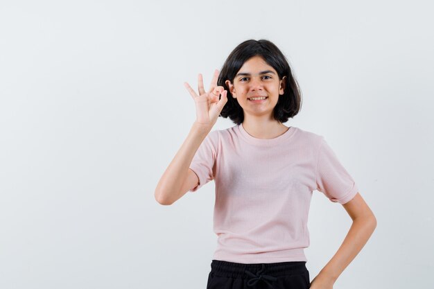 Jong meisje dat ok teken in roze t-shirt en zwarte broek toont en gelukkig kijkt