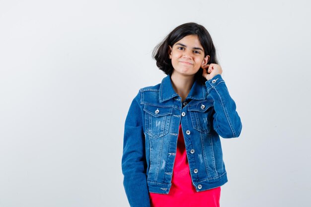 Jong meisje dat oor met vinger in rood t-shirt en jeansjasje uitrekt en vrolijk kijkt. vooraanzicht.