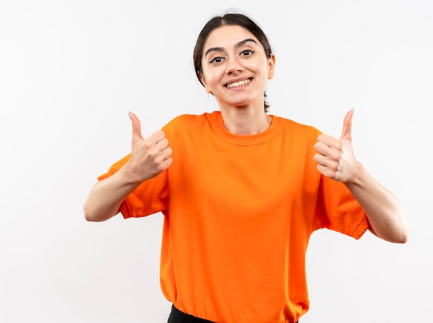 Jong meisje dat oranje t-shirt draagt dat met gelukkig gezicht glimlacht dat duimen toont die zich over witte muur bevinden