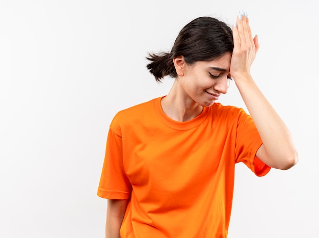 Jong meisje dat oranje t-shirt draagt dat met hand op haar hoofd voor fout kijkt die zich over witte muur bevindt