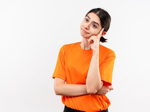 Jong meisje dat oranje t-shirt draagt dat opzij in verwarring kijkt wordt ontevreden die zich over witte muur bevindt