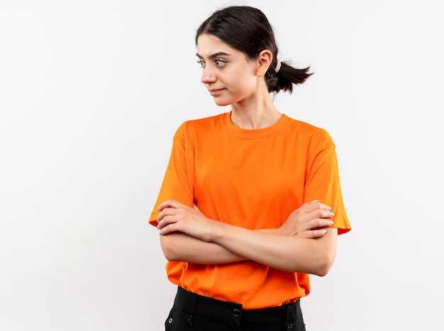 Jong meisje dat oranje t-shirt draagt dat opzij kijkt met ernstig gezicht dat wapens op borst kruist die zich over witte muur bevindt