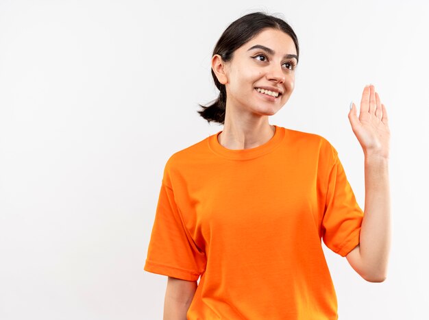 Jong meisje dat oranje t-shirt draagt die opzij glimlachend golvend met hand kijkt die zich over witte muur bevindt