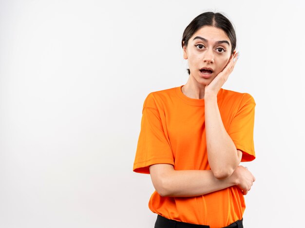 Jong meisje dat oranje t-shirt met hand op gezicht draagt dat zich over witte muur wordt verrast